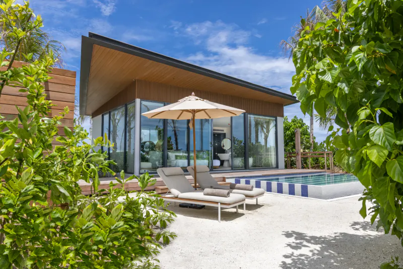 Modern beachfront villa with glass walls, beige umbrella, lounge chairs by infinity pool, surrounded by lush greenery under blue sky