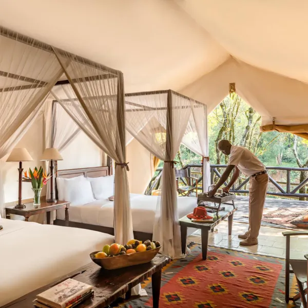 Luxurious tented suite at Fairmont Mara Safari Club with two canopy beds, staff serving tea, overlooking Kenyan savanna.