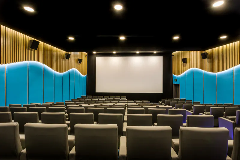 Modern cinema auditorium with rows of grey seats facing large blank screen, wood and blue wave walls, warm lighting.