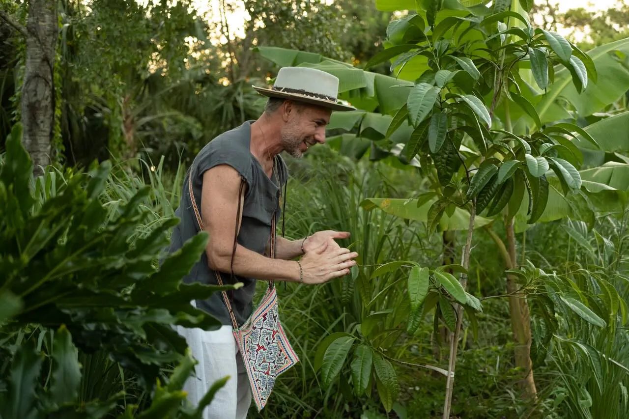Tony Cho claps hands smiling in tropical garden, wearing beige hat, tank top, white pants, and crossbody bag