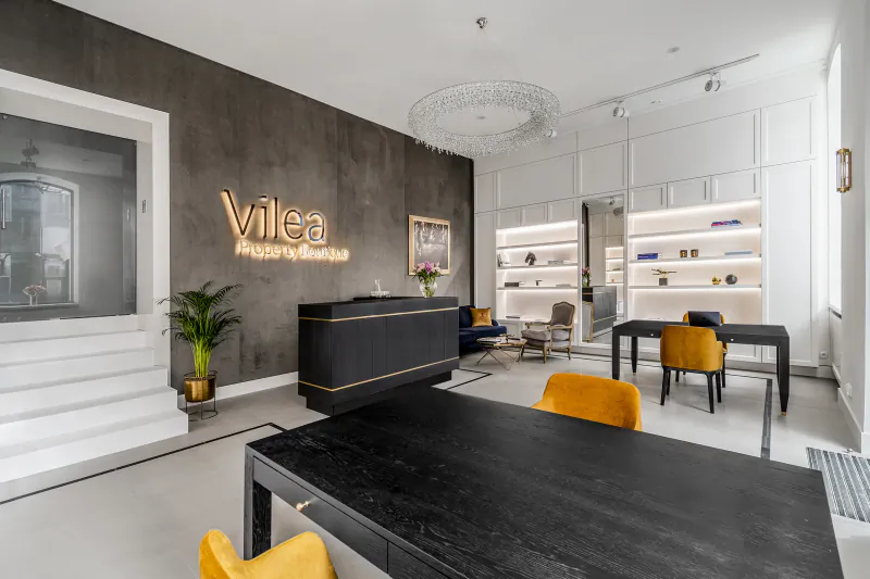 Modern Vilea Adrian office interior with neon 'Vilea' sign, dark gray wall, reception desk, orange chairs, bookshelves, chandelier, and plants.