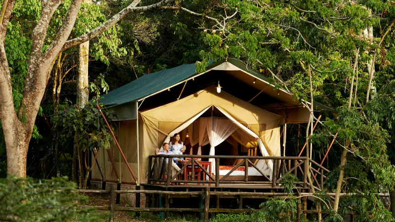 Luxury tented suite on raised deck at Fairmont Mara Safari Club, Kenya, with couple on balcony amid lush rainforest, overlooking Great Migration.