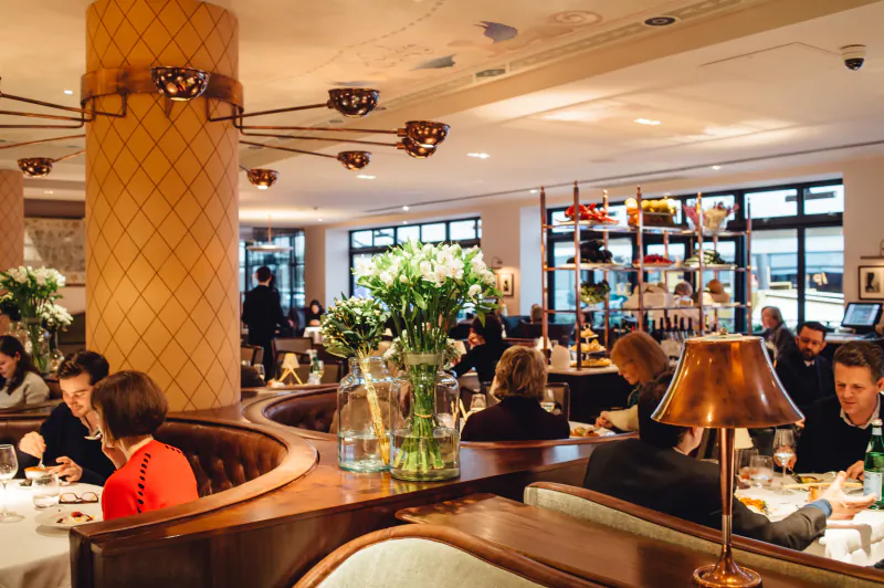 Elegant Sartoria Restaurant interior: patrons dining at curved wooden booths with large white flower arrangements, warm lighting, and ornate columns.