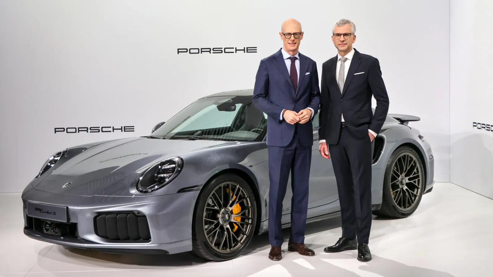 Two suited executives, one with glasses, stand beside a sleek gray Porsche 911 sports car in a studio.