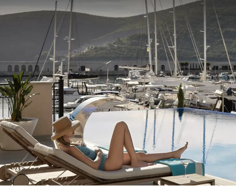 Woman in turquoise bikini and hat lounging on chaise by infinity pool overlooking marina and mountains