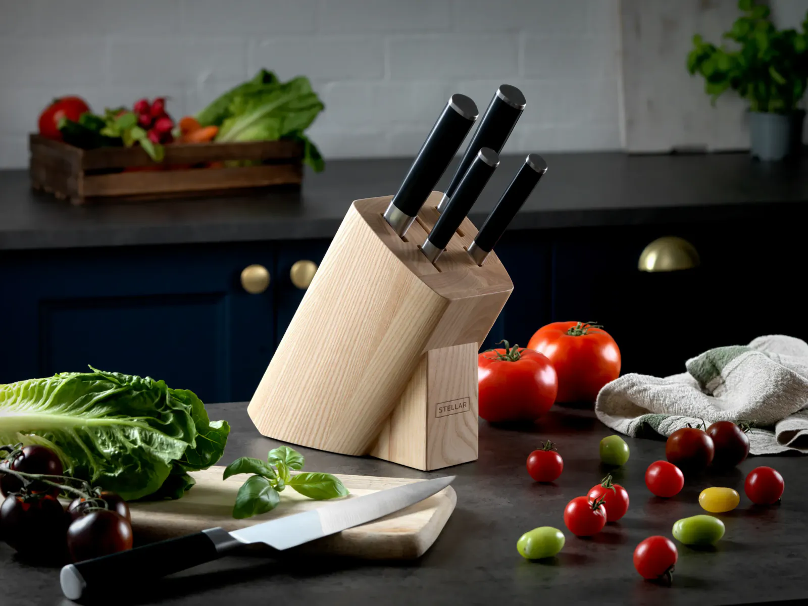 Wooden knife block with black-handled knives on kitchen counter amid fresh vegetables like tomatoes, lettuce, and herbs.