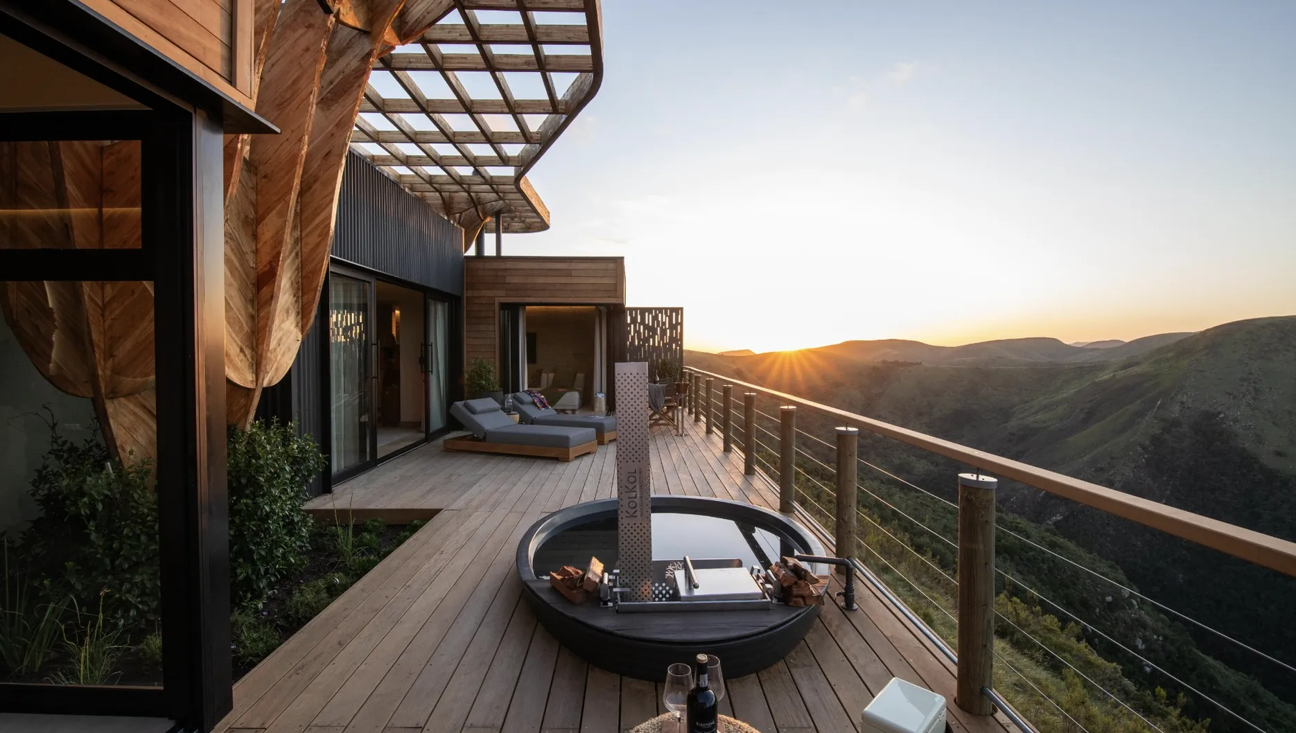 Luxury wooden deck with hot tub, fire pit, lounge seating, and sunset mountain views at Hiddn in Addo