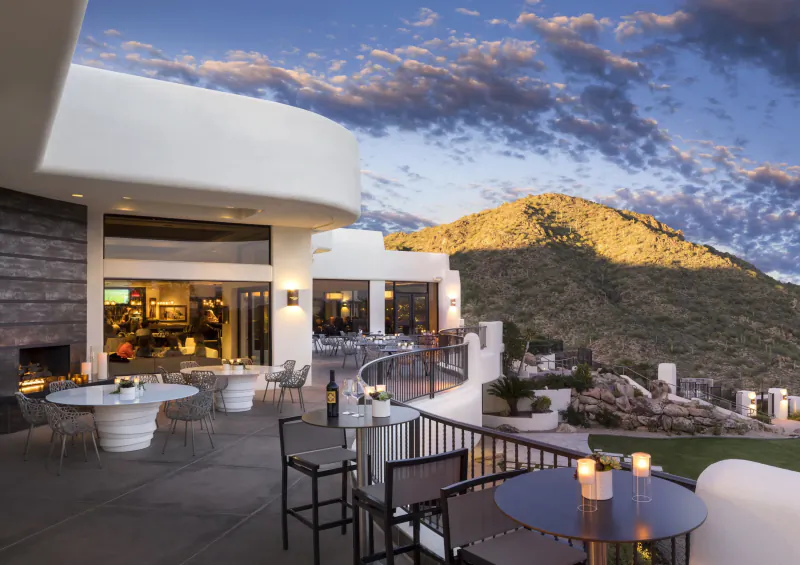Luxury outdoor terrace with white tables, chairs, candles at sunset, overlooking mountains and modern building entrance.