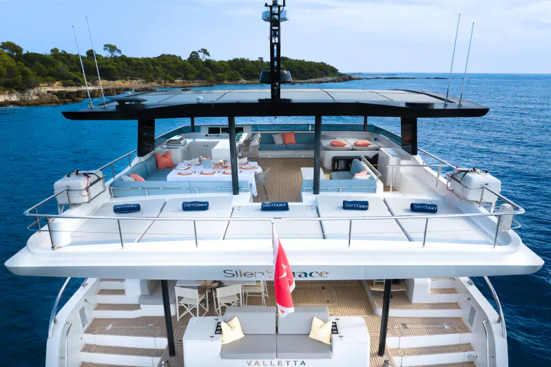 Front view of luxury yacht Vallatta on blue sea with lounge deck, cushions, furniture, and Singapore flag amid pines.