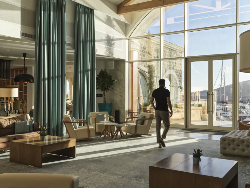 Man walking through elegant hotel lobby at The Chedi Luštica Bay Montenegro, with large windows overlooking marina.