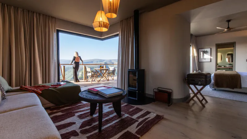 Cozy suite in Hiddn Addo with woman gazing from corner window at sunny mountain vista, lounge, bed, lamps.