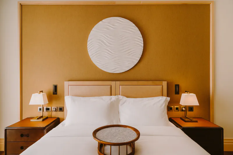 Luxurious hotel bed with white linens, wooden nightstands, lamps, and round wall art on tan wall