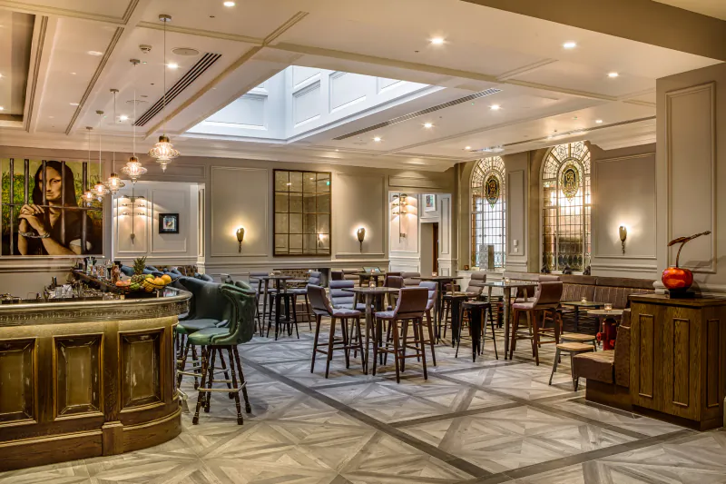 Courthouse Shoreditch elegant atrium restaurant with curved wooden bar, green stools, wooden tables, large portrait, and skylight.
