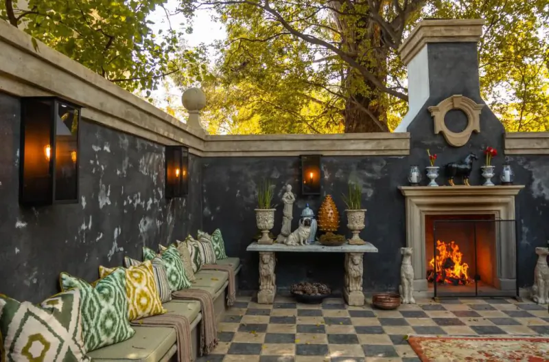Cozy outdoor patio with lit fireplace, green cushions on bench, statues, lanterns, trees, and checkered floor.