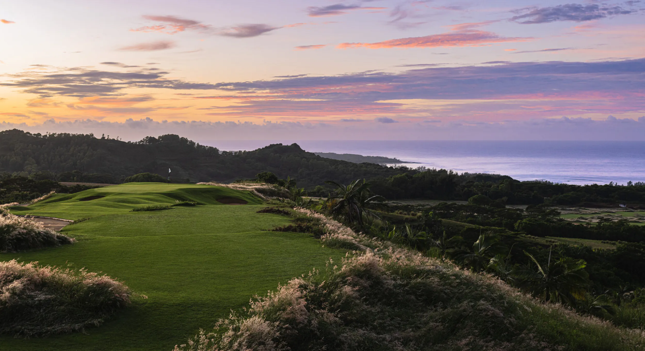 Sunset view of lush green golf course on coastal cliffs with ocean horizon and pink-purple sky.