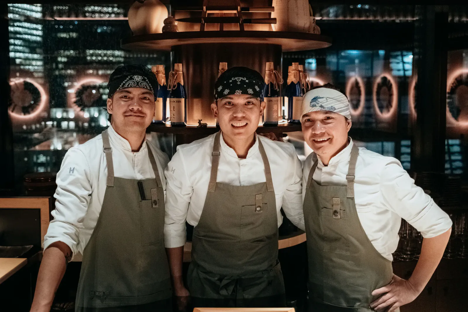 Three smiling chefs in white uniforms and green aprons pose in front of a glowing bar with city skyline at night.