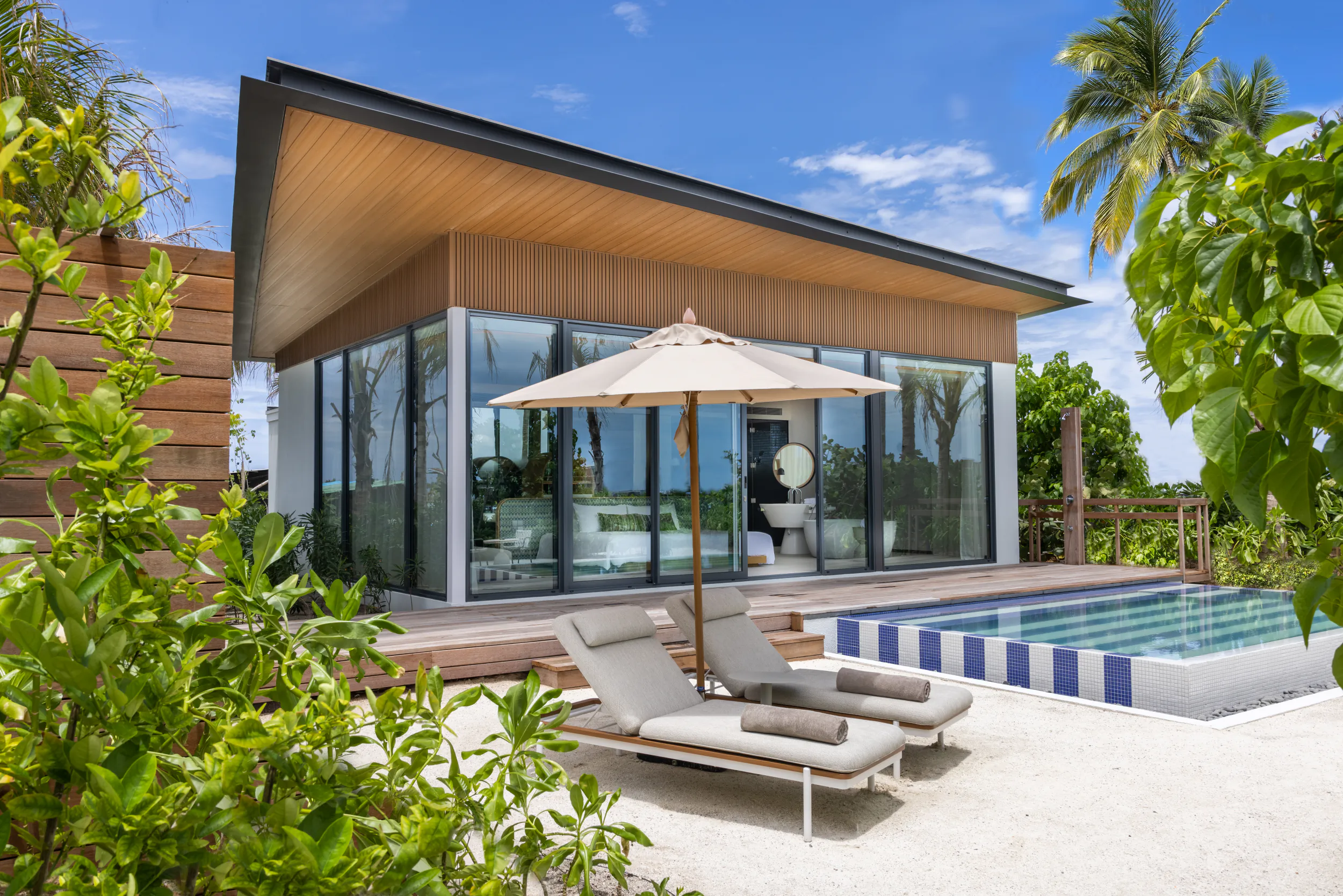 Modern wooden beachfront villa with infinity pool, lounge chairs, umbrella, and tropical palms under blue sky