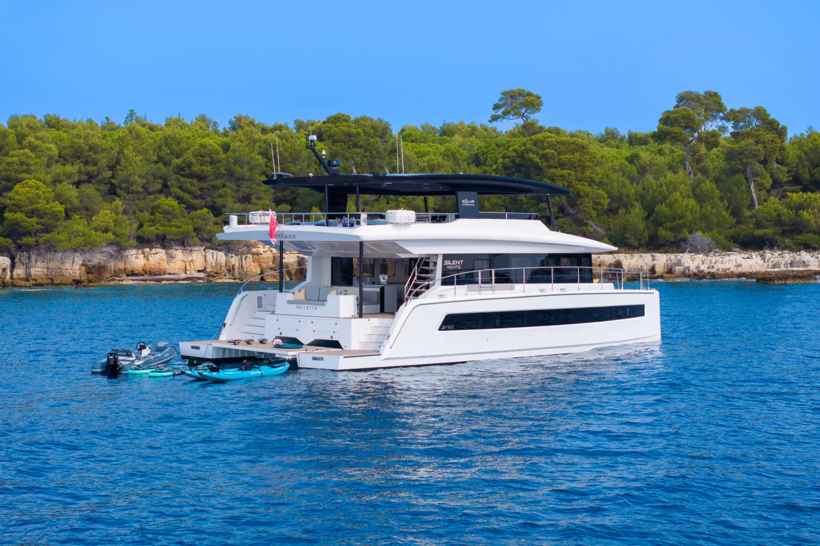 White luxury catamaran Silent Grace anchored in turquoise sea amid green pines and cliffs, with tender boat nearby.