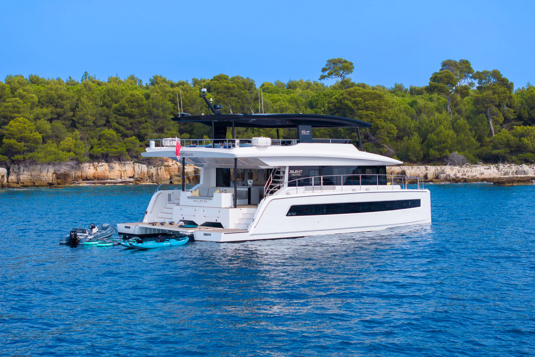 White luxury catamaran Silent Grace anchored in turquoise sea amid green pines and cliffs, with tender boat nearby.