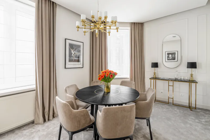 Elegant dining room with black round table, beige chairs, gold chandelier, red flowers, curtains, and console table.