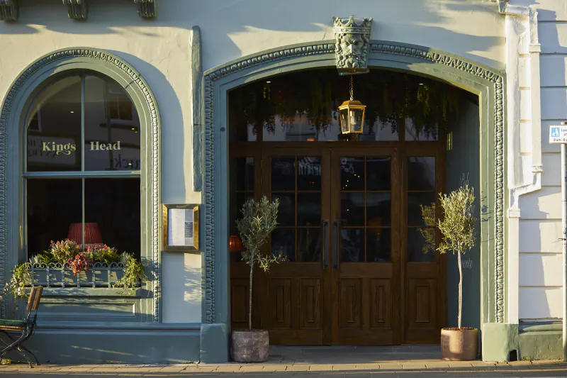 Kings Head inn facade in Cirencester: arched green door with lion gargoyle, lantern, olive trees, flowers in window.