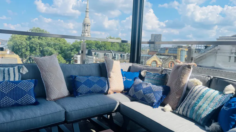 Cozy rooftop seating at Courthouse Shoreditch with blue, beige pillows, city skyline, spire, trees under blue sky.