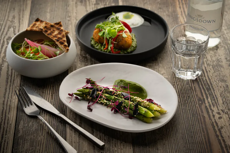 Assortment of dishes on wooden table: asparagus with purple tips and dip, curried fritter with greens, salad, crackers, water glass