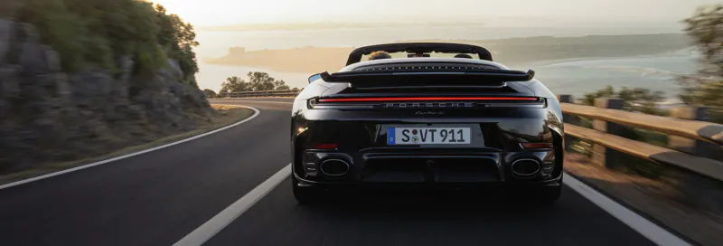 Rear view of black Porsche 911 Cabriolet with plate 'EU 57071' driving coastal road at sunset amid cliffs and sea.