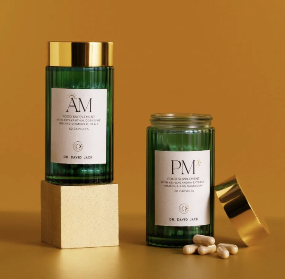 Two green glass supplement bottles labeled AM and PM by Dr. David Jack on a yellow background, with capsules spilled beside the open PM bottle