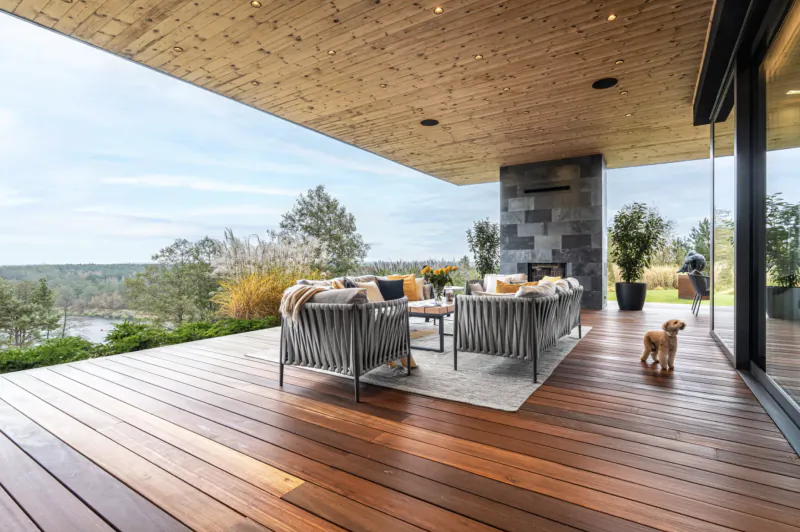 Modern outdoor deck with woven gray lounge chairs, pillows, small dog, overlooking lake and trees under wooden ceiling