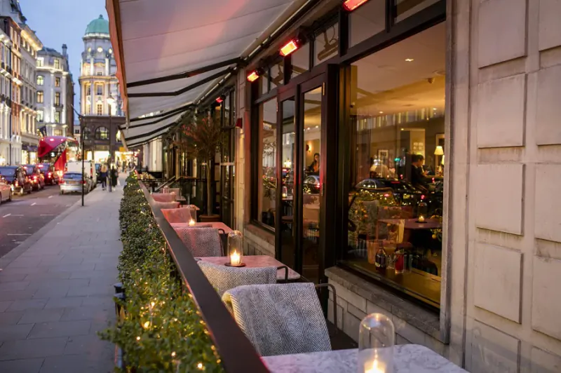Sartoria Restaurant on Savile Row, London: elegant outdoor terrace with candlelit tables, heaters, greenery, and city buildings at dusk.