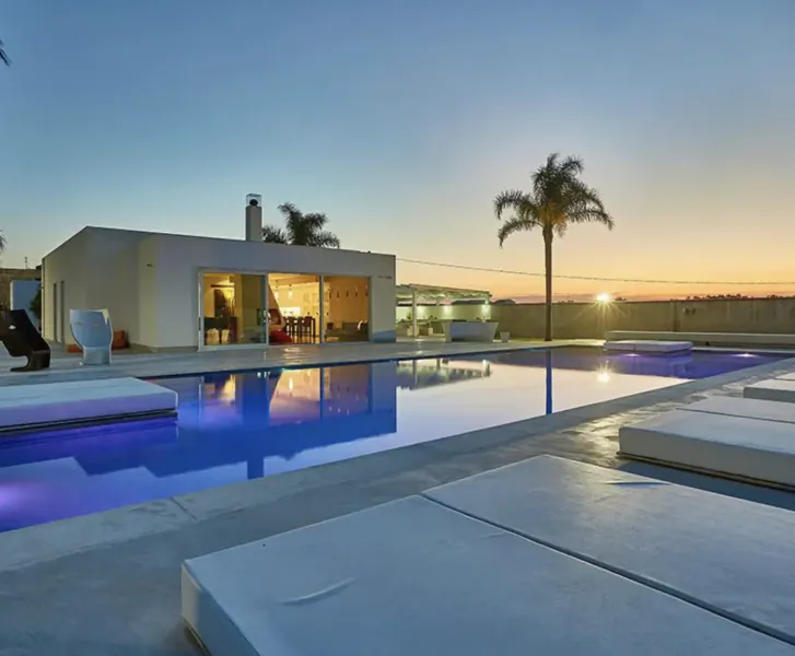 Modern white villa with infinity pool, white loungers, and palm trees at sunset.
