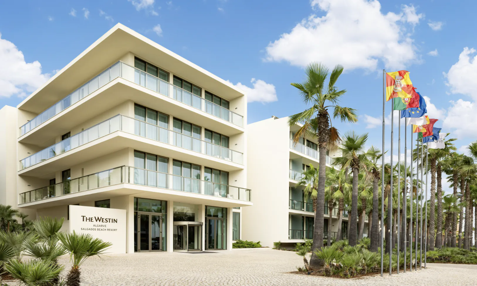 The Westin Sallgados Beach Resort, modern white multi-story building with glass balconies, palms, flags, and entrance in Algarve, Portugal