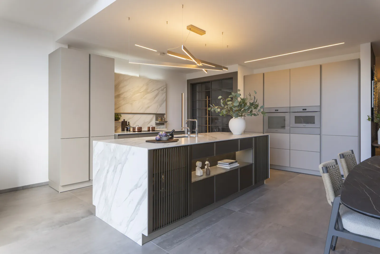 Modern kitchen with white marble island, dark wood cabinetry, built-in ovens, and dining table in a bright showroom