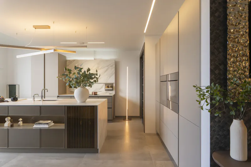 Modern kitchen showroom with large island, white vase with greenery, tall floor lamp, and built-in ovens
