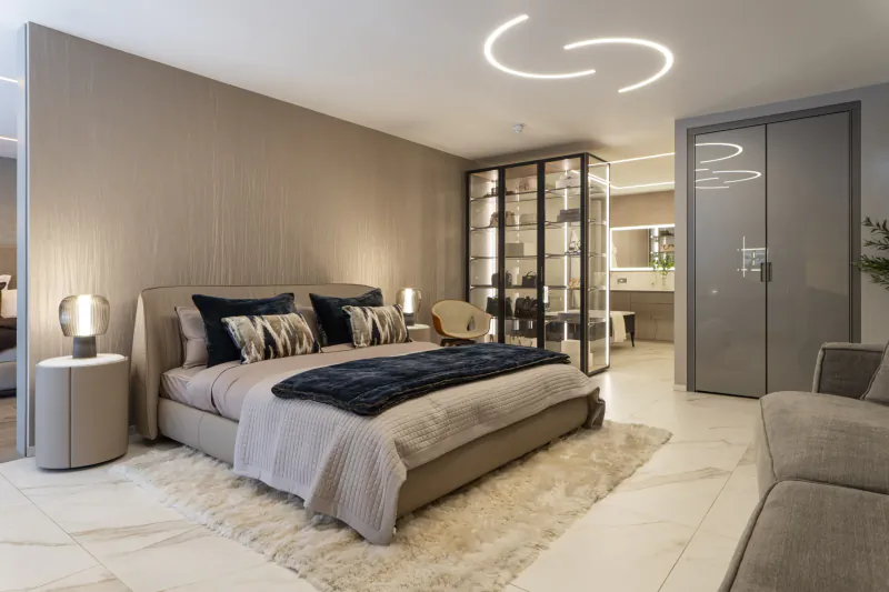 Modern bedroom with beige upholstered bed, navy pillows, glass display cabinet, and curved ceiling lights