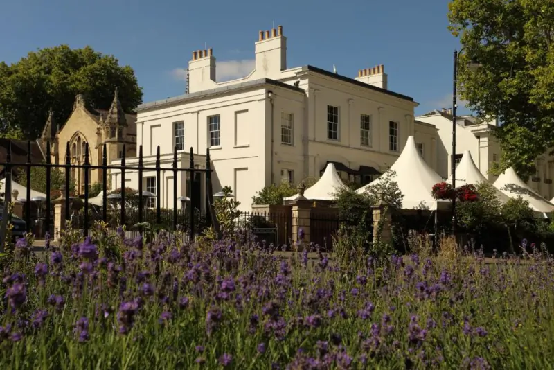 White Georgian mansion with chapel beside it, white marquees, purple lavender field, and trees under blue sky