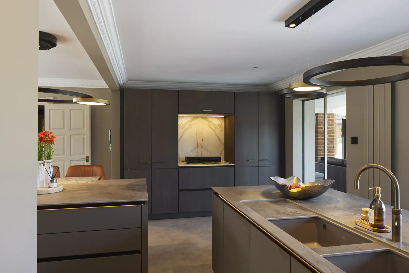 Modern grey kitchen with central island, fruit bowl, sink, pendant lights, and orange flowers by door.