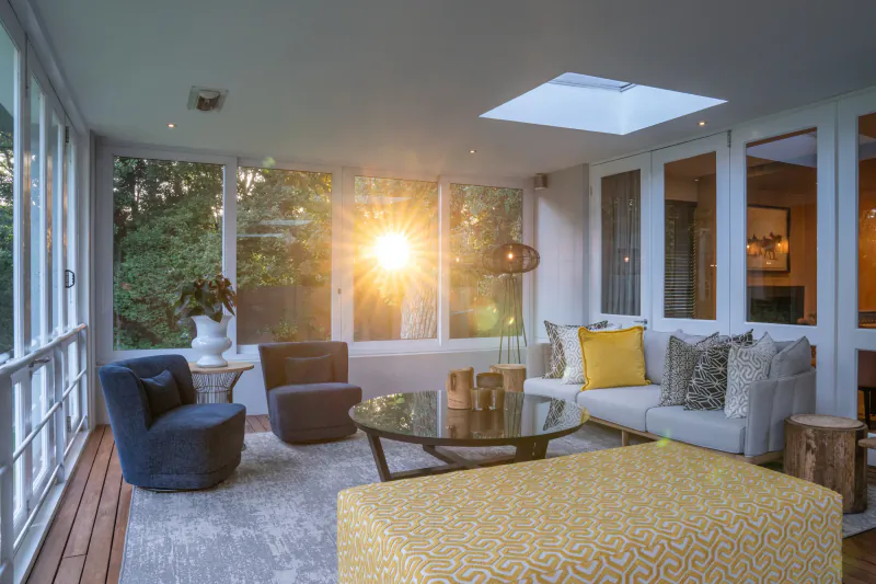 Sunlit sunroom in luxury villa with gray rug, navy armchairs, yellow ottoman, glass coffee table, and trees outside.