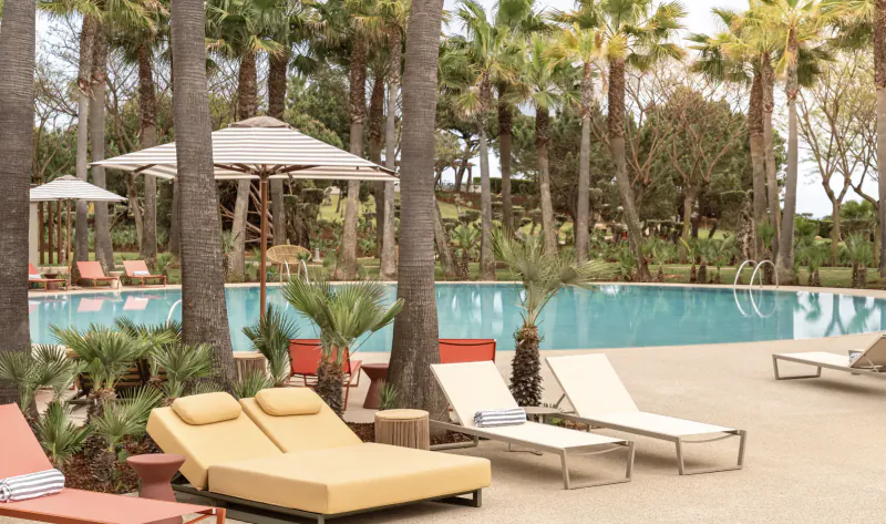 Luxury poolside at Kimpton Portugal resort with yellow and white lounge chairs, umbrellas, and palm trees.