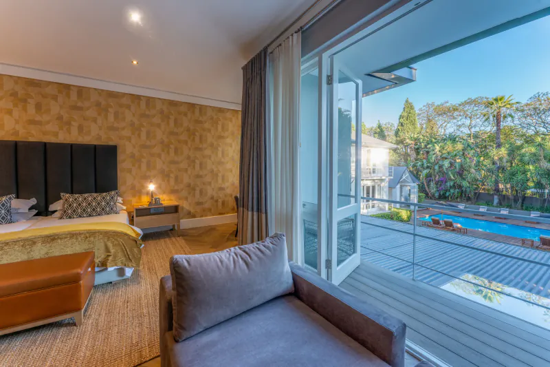 Luxury hotel bedroom with king bed, armchair, and open doors to balcony overlooking pool and gardens.