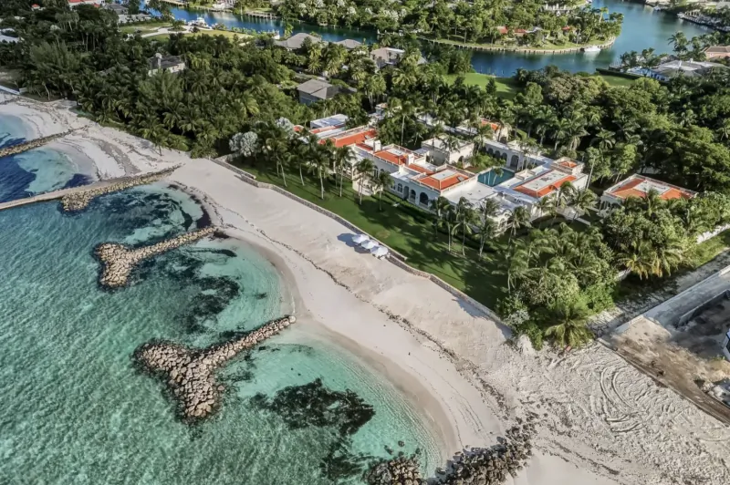 Aerial view of luxurious red-tiled beachfront mansion in Bahamas, surrounded by turquoise waters, breakwaters, palms, and lawns.