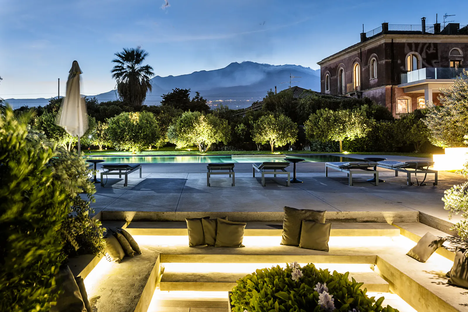 Twilight poolside at Zash Country Boutique Hotel & Spa with lit gardens, olive trees, palm, mountains, and historic villa.