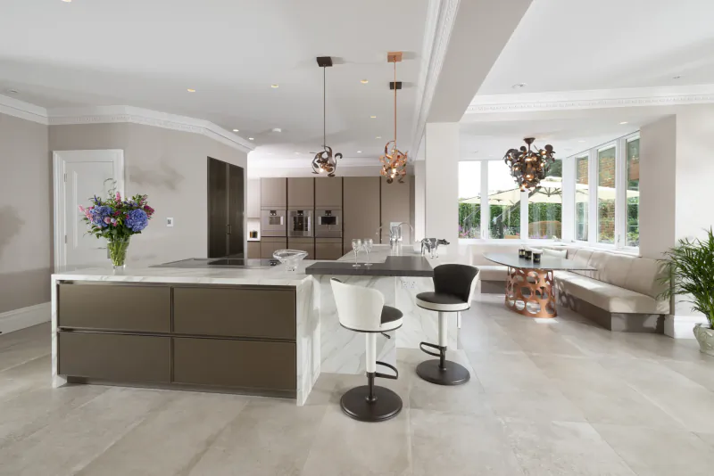 Modern kitchen with white marble island, brown cabinets, white bar stools, chandelier lights, flowers, and window view