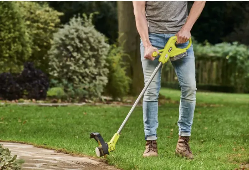 Person using a Ryobi cordless grass trimmer on a lawn near a stone path