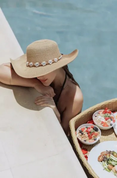 Woman in straw hat resting by pool with breakfast tray of yogurt bowls and salad