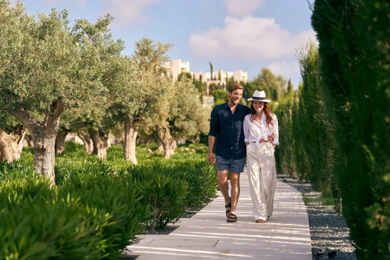 A man and woman walking together on a sunny garden path lined with olive trees and green hedges