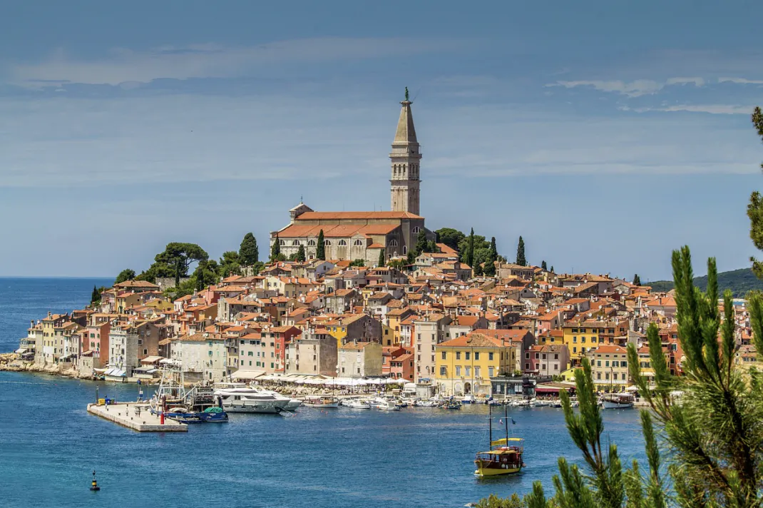 Aerial view of Rovinj, Croatia: hilltop church with bell tower overlooking colorful terracotta rooftops, marina with boats, and Adriatic Sea.