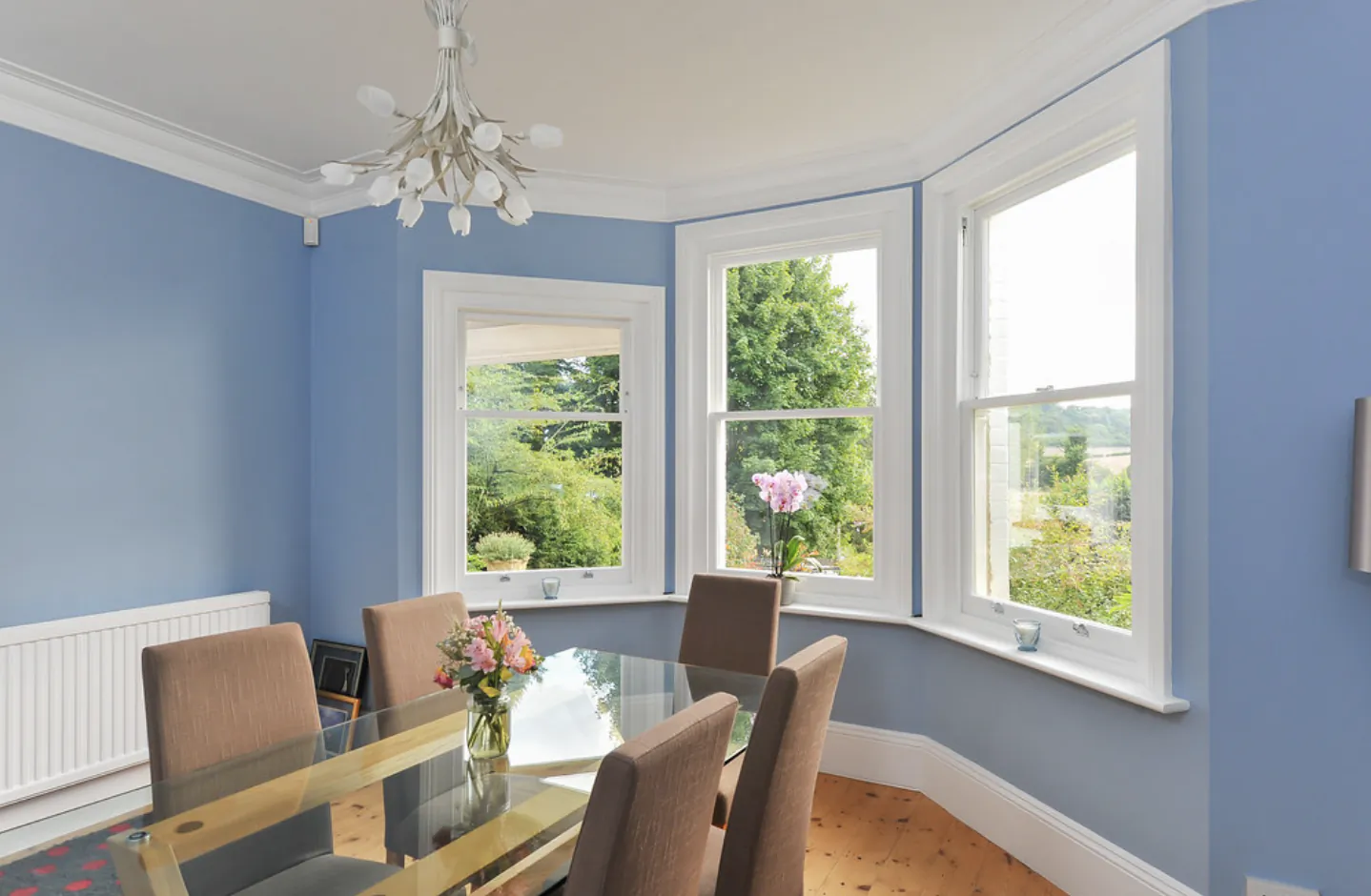 Bright dining room with blue walls, glass table, four chairs, and three large sash windows overlooking greenery