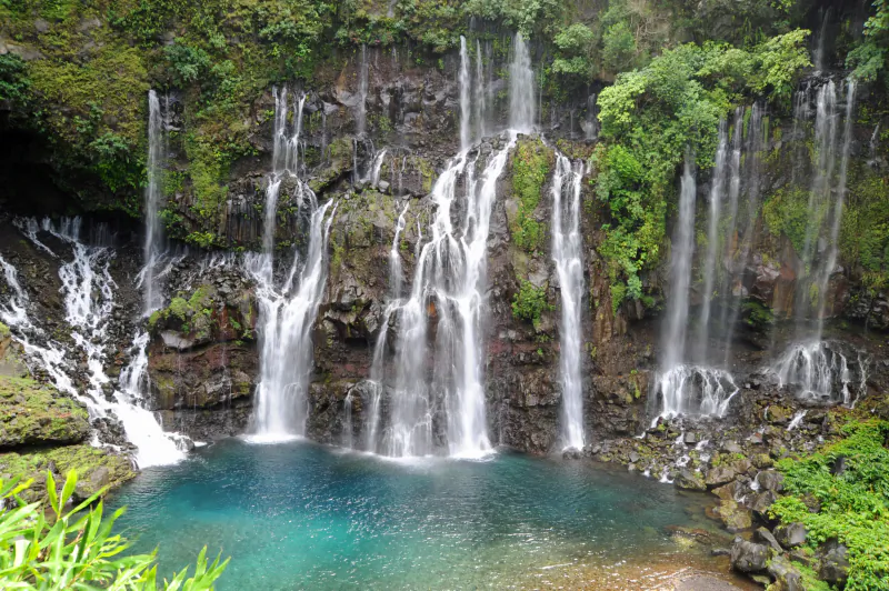 Multiple waterfalls cascading down a lush green rocky cliff into a turquoise pool